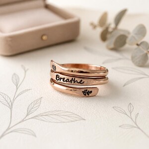May include: A rose gold spiral ring with the word "Breathe" engraved in black script. The ring features leaf designs and is displayed near a ring box and decorative foliage.