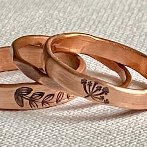 May include: Three copper rings with floral designs. The rings are a warm copper color and feature etched botanical patterns. The rings are stacked together, showcasing the intricate details of the engraved leaves and flowers.