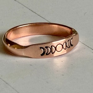 May include: A rose gold band ring with a moon phase design engraved on the top. The design features a full moon, crescent moons, and a half moon.