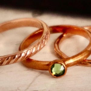 May include: Three gold rings with a green gemstone set in the center ring. The rings are stacked on top of each other.