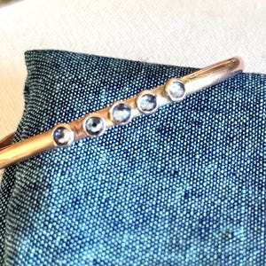 May include: A rose gold cuff bracelet with six round, light blue gemstones. The bracelet is displayed on a textured blue fabric, showcasing its polished finish and the sparkle of the stones. A simple and elegant piece of jewelry.