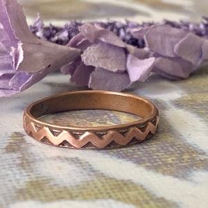 May include: A thin rose gold band ring with a zig-zag pattern around the edge.