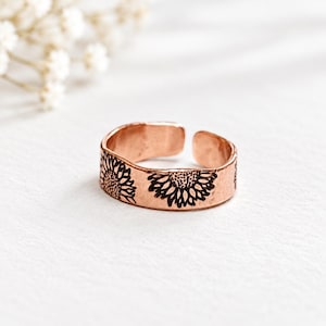 May include: A copper-toned metal ring featuring an open-ended band design. The exterior surface of the ring is engraved with a repeating black sunflower pattern that wraps around the circumference of the band, showcasing detailed petal textures.
