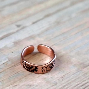 May include: A copper-colored adjustable ring with a floral design. The ring features a band with etched black sunflowers. The open design allows for size adjustment. The ring is displayed on a light wooden surface.
