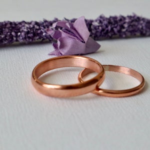 May include: Two simple copper wedding bands, one slightly larger than the other, are lying on a white surface. The bands are smooth and shiny, with a warm copper color.