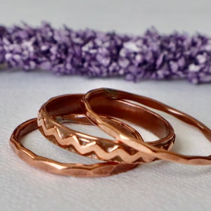 May include: Three copper rings with unique textures and designs. One ring features a zigzag pattern, another has a hammered texture, and the third is smooth. The rings are displayed against a soft, blurred background with purple accents.