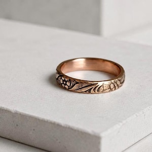 May include: A rose gold ring with a floral pattern. The ring features detailed carvings of flowers and leaves around the band. The ring is displayed on a white surface.