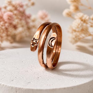 May include: A copper-colored spiral ring with a star and crescent moon design. The ring wraps around the finger with a celestial theme. The star and moon are etched into the metal, adding detail to the jewelry.
