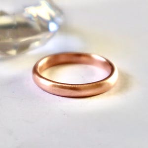 May include: A rose gold ring with a smooth, rounded design. The ring is set against a white background, with a blurred, faceted object in the background. The ring's warm color and simple design suggest a piece of jewelry.