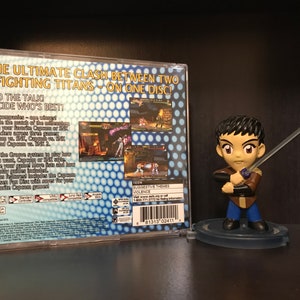 May include: The back of a video game case for Capcom vs. SNK 2. The case is blue and white with a grid pattern. The text on the case reads "The Ultimate Clash Between Two Fighting Titans - On One Disc!" and "End the Talk! Decide Who's Best!"
