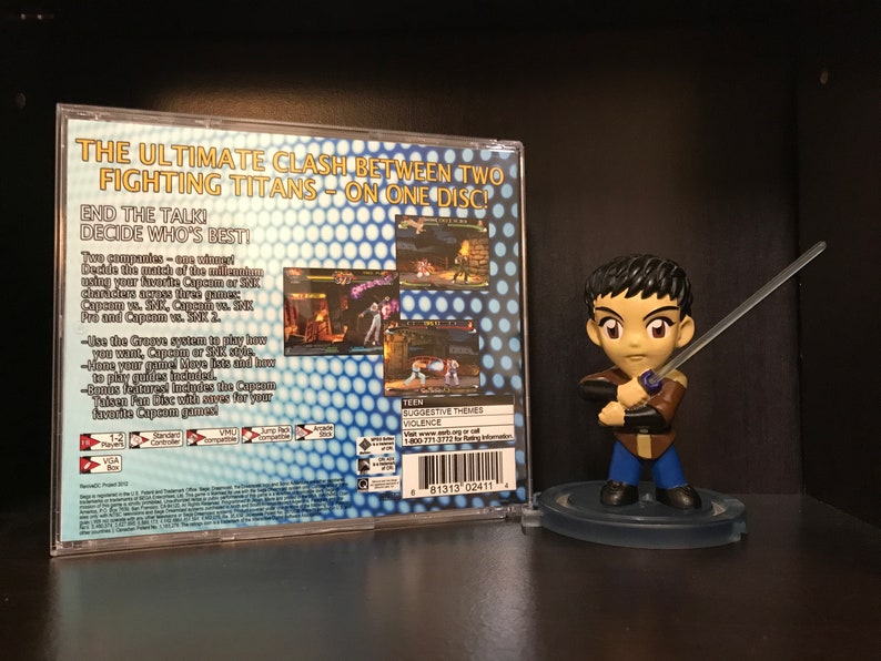 May include: The back of a video game case for Capcom vs. SNK 2. The case is blue and white with a grid pattern. The text on the case reads "The Ultimate Clash Between Two Fighting Titans - On One Disc!" and "End the Talk! Decide Who's Best!"