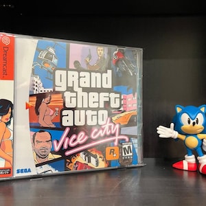May include: A Sega Dreamcast game case for Grand Theft Auto: Vice City. The case is blue and white with a picture of a woman in a bikini, a man in a suit, and a car. The game is rated R and M for Mature.