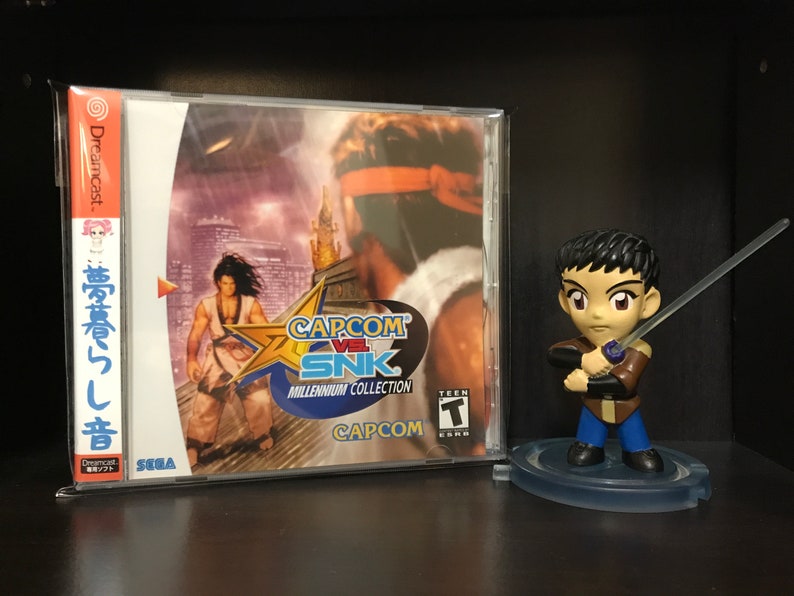 May include: A Dreamcast video game case for Capcom vs SNK: Millennium Collection. The case is silver with a red and white label. The label features a red star with the game title in white text. The game is rated Teen by the ESRB. A small figurine of a character from the game is standing on a clear plastic base.