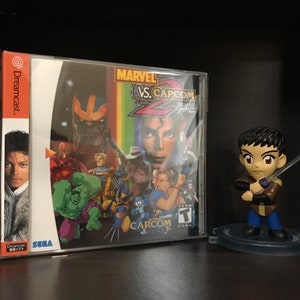 May include: A Sega Dreamcast game case for Marvel vs. Capcom 2: New Age of Heroes. The case is in a plastic sleeve and features a colorful image of characters from the game, including Thanos, Hulk, Wolverine, and Ryu. The game is rated T for Teen by the ESRB.