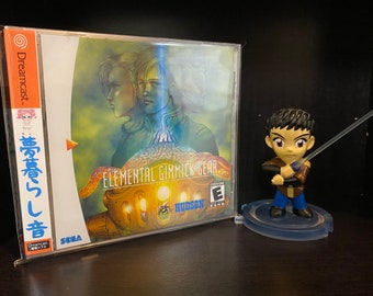 May include: A Sega Dreamcast game case with a blue and yellow cover featuring the text "Elemental Gimmick Gear" and a character with a sword. The case is in a plastic sleeve. A small figurine of a character with a sword is in the foreground.