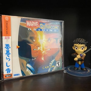 May include: A Dreamcast video game case with a blue and orange cover featuring the text "MARVEL VS CAPCOM ORIGINS". The game case is in a clear plastic protector. A small figurine of a character holding a sword is in the foreground.