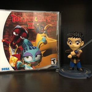 May include: A Sega Dreamcast game case for ToeJam & Earl III, featuring the three main characters on a brick wall background. The game case is next to a small figurine of a young man holding a sword.