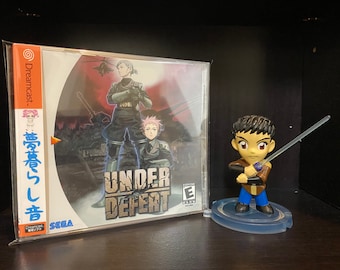 May include: A Sega Dreamcast game case for Under Defeat, a military-themed action game. The case is in a plastic sleeve and features a red, orange, and yellow color scheme. The game title is printed in black and white on the front of the case. The case is next to a small figurine of a character from the game, holding a sword.