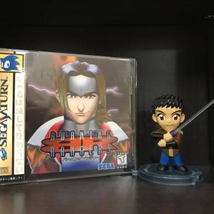 May include: A Sega Saturn video game case with a blue and gold design. The game is called "Burning Rangers" and features a character with red and blue clothing. A small figurine of a character with a sword is in the foreground.