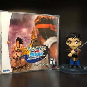 May include: A Sega Dreamcast game case for Capcom vs SNK: Millennium Collection. The case is white with a blue and yellow star on the front. The game features a character in a white and blue outfit standing on a rooftop. The case also features a Teen rating sticker. A small figurine of a character from the game is standing on a clear base.