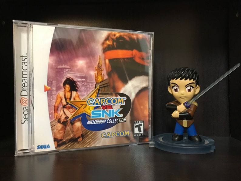 May include: A Sega Dreamcast game case for Capcom vs SNK: Millennium Collection. The case is white with a blue and yellow star on the front. The game features a character in a white and blue outfit standing on a rooftop. The case also features a Teen rating sticker. A small figurine of a character from the game is standing on a clear base.