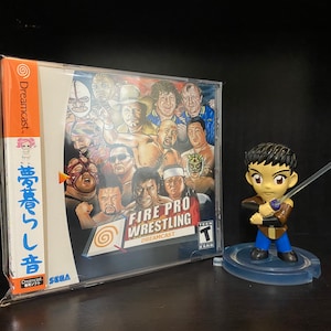 May include: A Dreamcast video game case for Fire Pro Wrestling, featuring a collage of wrestlers. A small anime-style figure with a sword stands to the right. The game case is in a protective sleeve.