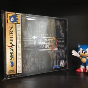 May include: A Sega Saturn video game case with a black and white image of a futuristic city on the front. The game title is "Blast Wind" in white text on a red and yellow background. The game is rated E for Everyone by the ESRB.