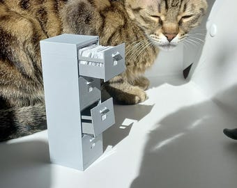 Puede incluir: Un archivador en miniatura de color gris, impreso en 3D, con cuatro cajones, uno de los cuales está abierto. Un gato atigrado está tumbado sobre una superficie blanca junto al archivador.