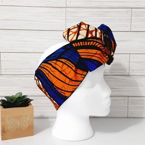 May include: A vibrant, patterned headband in orange, blue, and brown, displayed on a white mannequin head. The fabric features a bold, abstract design. The headband is a stylish accessory.