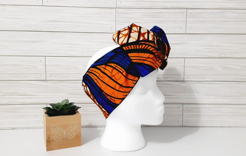 May include: A vibrant, patterned headband in orange, blue, and brown, displayed on a white mannequin head. The fabric features a bold, abstract design. The headband is a stylish accessory.