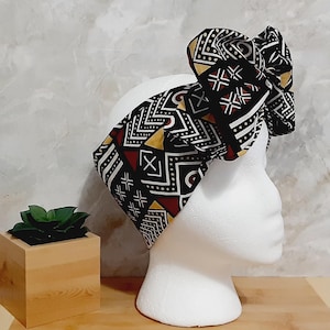 May include: A black, white, and red patterned headband with a large bow on top. The headband is displayed on a white mannequin head. The design features geometric shapes and lines. The background includes a small wooden planter with a green plant.