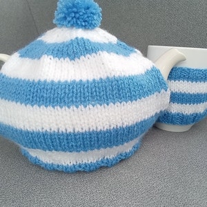 May include: A knitted tea cosy and matching mug cosy set. The cosy set features alternating horizontal stripes of light blue and white. The tea cosy has a blue pom-pom on top. The set is designed to keep tea warm.