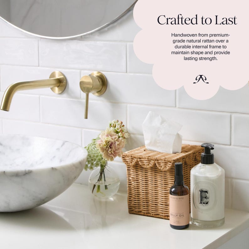 May include: A bathroom scene featuring a marble sink, gold faucet, and a woven rattan tissue box. The countertop also holds a small glass vase with flowers, a bottle of oil, and a white bottle with a black pump. The text reads "Crafted to Last".