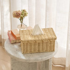 May include: A rectangular woven wicker tissue box in a light beige color. The tissue box has a scalloped edge design and a rectangular opening for tissue dispensing. A stack of tissues is visible. The tissue box sits on a marble table.
