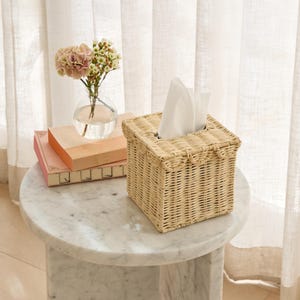 May include: A square, woven wicker tissue box in a natural beige color. White tissues are pulled from the top opening. The box sits on a marble-topped side table, next to a small glass vase with pink and white flowers and two books.