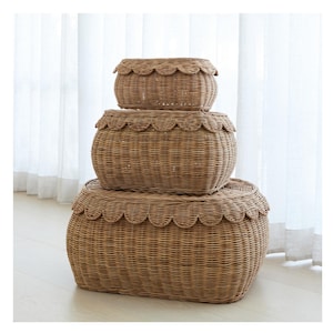 May include: Three woven wicker storage baskets stacked on top of each other. The baskets are oval-shaped and have a scalloped edge design. The baskets are a light brown color and are sitting on a light-colored wooden floor.