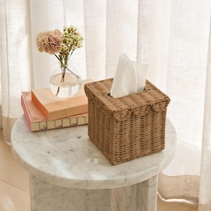May include: A square, woven wicker tissue box in a light brown hue. White tissues are visible at the top. The box sits on a white marble side table, next to a small vase with pink and white flowers and two books.