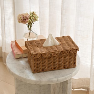 May include: A rectangular, woven wicker tissue box in a warm brown hue. The box features a scalloped edge design and a central opening for tissue dispensing. A small stack of tissues protrudes from the top. The box sits on a marble-topped side table.