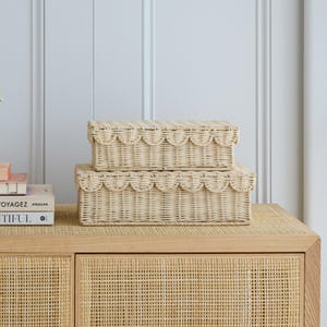 May include: Two woven wicker storage boxes with scalloped edges, one stacked on top of the other. The boxes are a light beige color and sit on a light wood surface. The background features a white wall and a wooden cabinet.