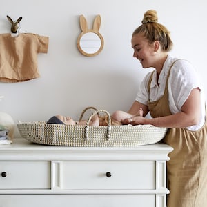 May include: A woven wicker baby changing basket with handles, placed on a white dresser. A baby lies inside. A wooden bunny mirror and a small tan shirt hang on the wall above. The room has a neutral, natural aesthetic.
