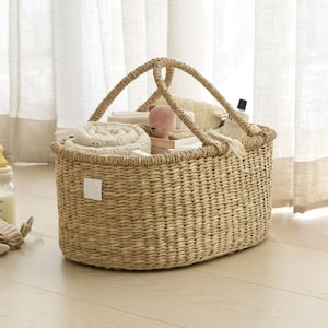 May include: A woven wicker basket with two arched handles, filled with baby items. The basket contains rolled blankets, a small stuffed animal, and bottles. The basket is light brown and sits on a light wood floor.
