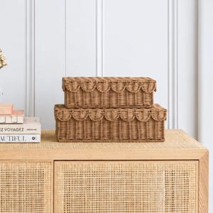 May include: Two rectangular, light brown woven wicker storage boxes with lids are stacked on top of each other. Each box features a decorative scalloped edge detail along the top rim. The boxes have a natural, textured finish.