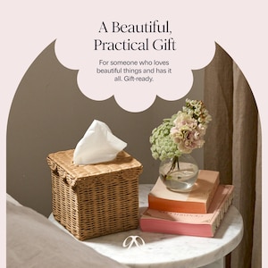 May include: A woven wicker tissue box with a white tissue peeking out. The box is square and sits on a marble-topped table next to a small glass vase with flowers and a stack of books. The text reads: "A Beautiful, Practical Gift."