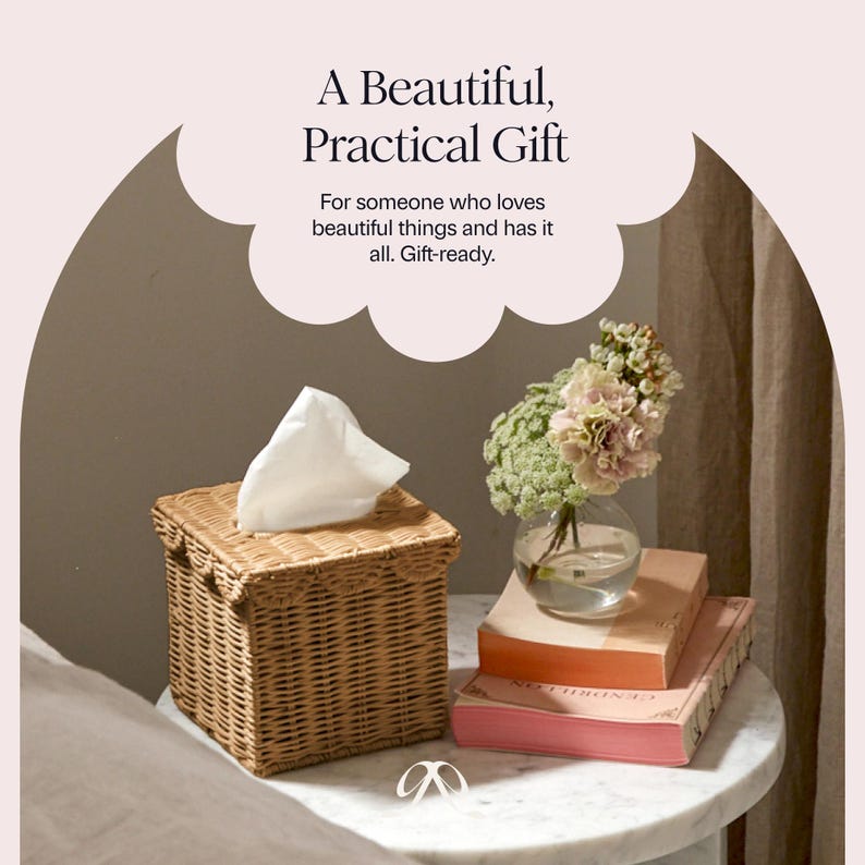May include: A woven wicker tissue box with a white tissue peeking out. The box is square and sits on a marble-topped table next to a small glass vase with flowers and a stack of books. The text reads: "A Beautiful, Practical Gift."