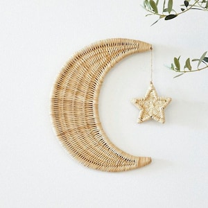 May include: A woven rattan crescent moon wall hanging with a hanging star. The moon and star are made of natural-colored rattan and are suspended against a white background. The star hangs from a string attached to the moon.