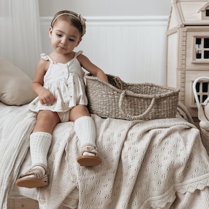 May include: A young child sits on a bed with a woven basket. The child wears a cream-colored dress, white knee-high socks, and sandals. A cream-colored blanket with a lace trim covers the bed. A dollhouse is in the background.