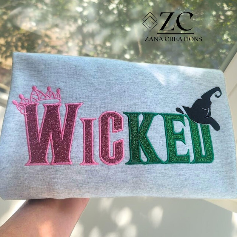 May include: A light gray sweatshirt with the word "Wicked" in pink and green glitter lettering. A pink crown is above the "W" and a black witch hat is above the "E". The text "Zana Creations" is in the upper right corner.