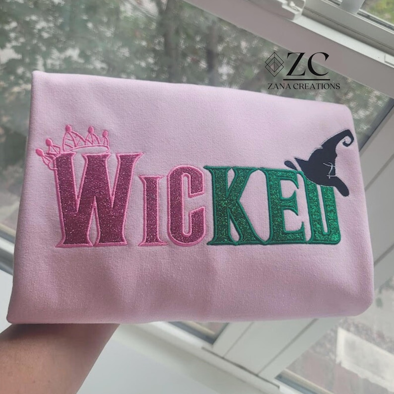 May include: A light pink textile with the word "Wicked" in glittery pink and green letters. A pink crown is above the "W" and a black witch hat is next to the "E". The text "ZANA CREATIONS" is in the upper right corner.