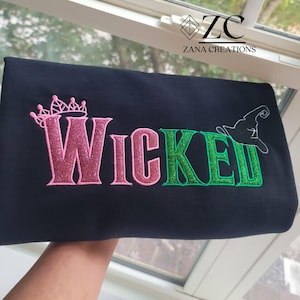 May include: Black fabric with the word "Wicked" embroidered in pink and green glitter thread. A pink crown is above the "W" and a witch hat is above the "E". The text "Zana Creations" is in the upper right corner.