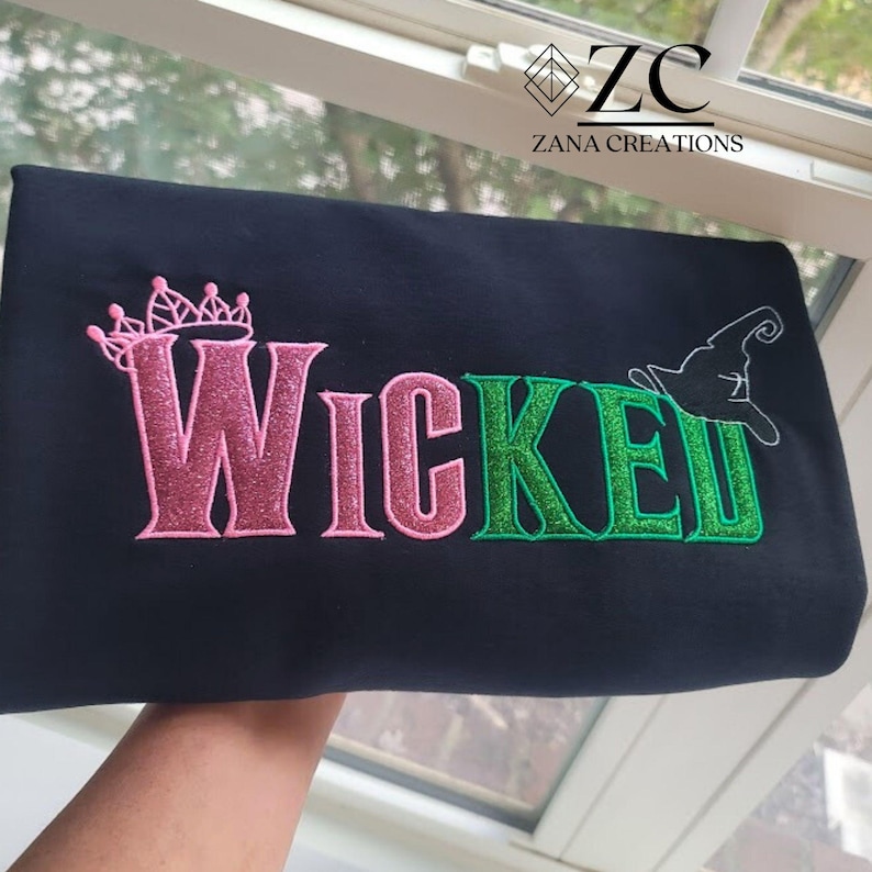 May include: Black fabric with the word "Wicked" embroidered in pink and green glitter thread. A pink crown is above the "W" and a witch hat is above the "E". The text "Zana Creations" is in the upper right corner.